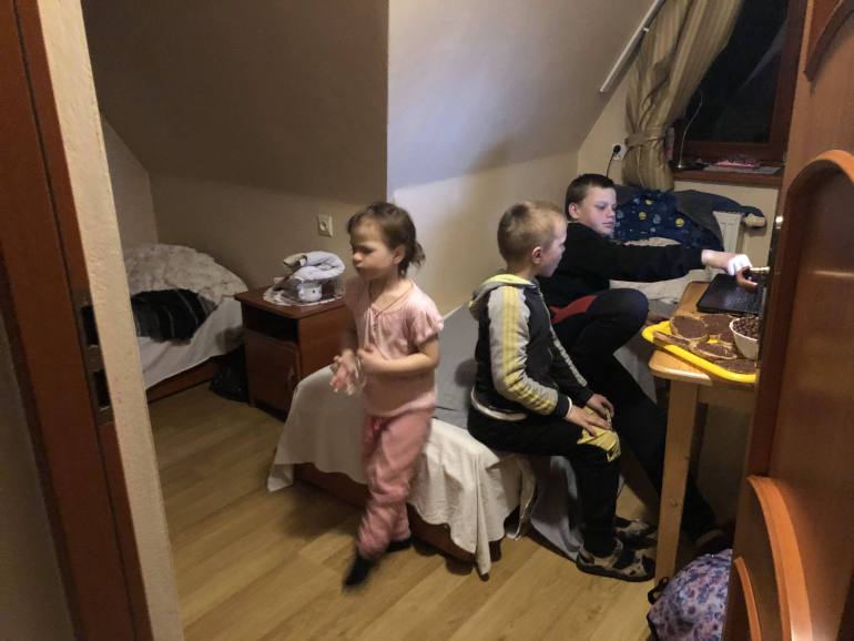 A photo of three children sitting in a room. one is on the computer, the other is sitting next to him and the third child is standing behind them.