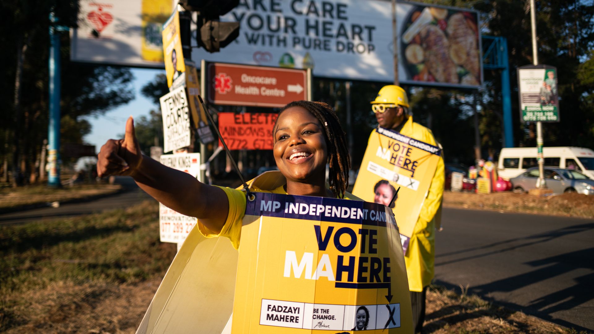 A photo of a woman wearing a yellow "Vote Mahre" sign and a man standing in the back wearing yellow clothes and holding a "Vote Mahre" sign.
