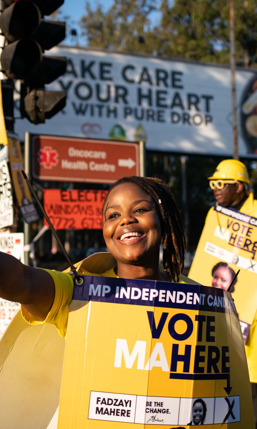 A photo of a woman wearing a yellow "Vote Mahre" sign and a man standing in the back wearing yellow clothes and holding a "Vote Mahre" sign.