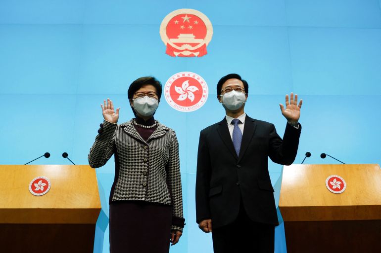 Carrie Lam (left) and John Lee (right) wave stiffly as they stand between lecterns in front of a bright blue wall with the Hong Kong and China plaques