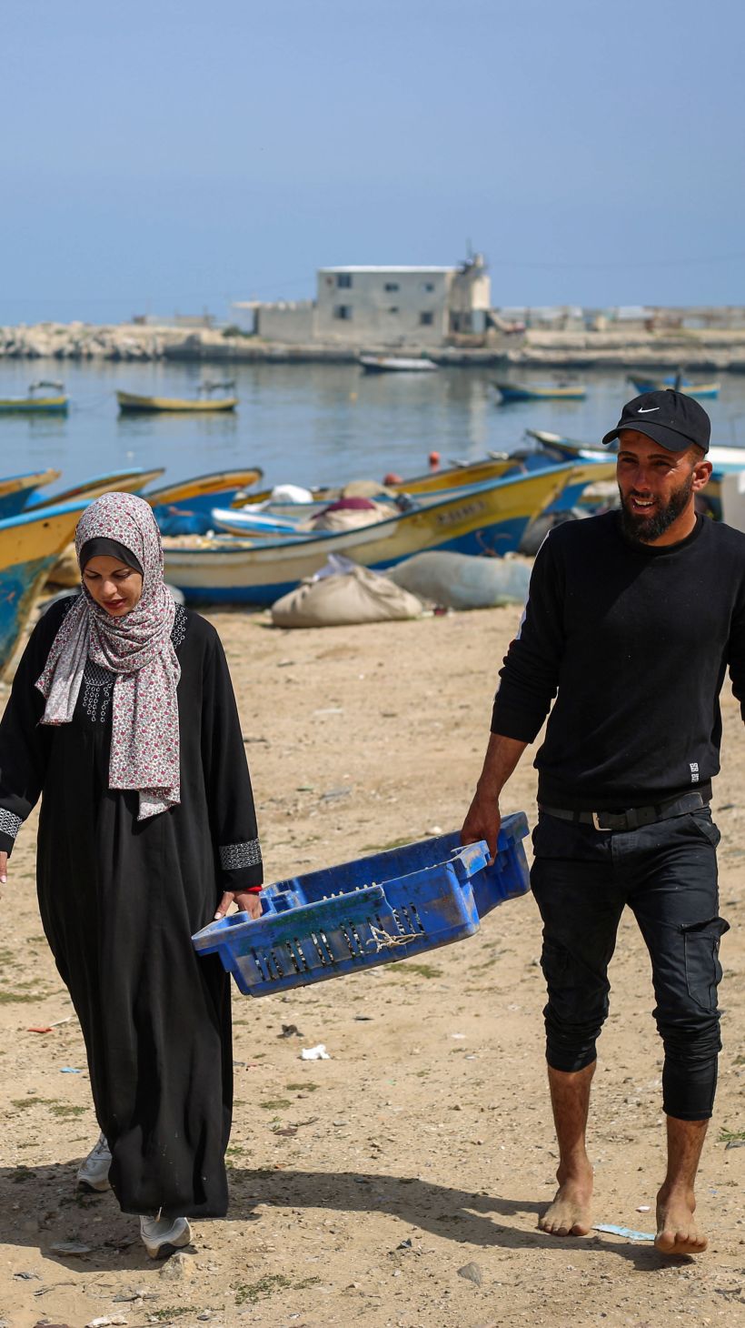 Khadr and Madleine walk on the beach with a blue basket of fish between them