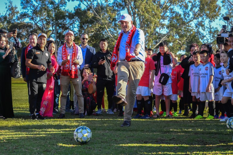 Scott Morrison in his customary beiuge chinos and blue shirt kicks a football towards the camera as children and adults from the Vietnamese community watch