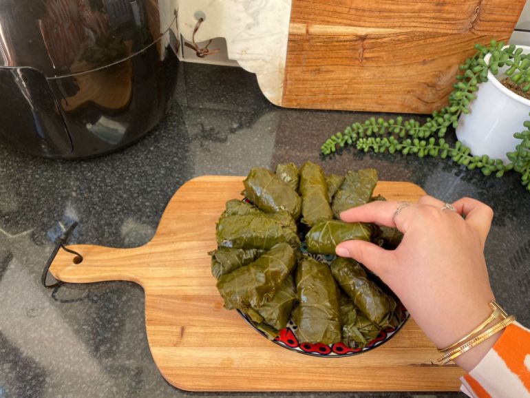 A hand is seen stacking rolled stuffed grape leaves on a plate