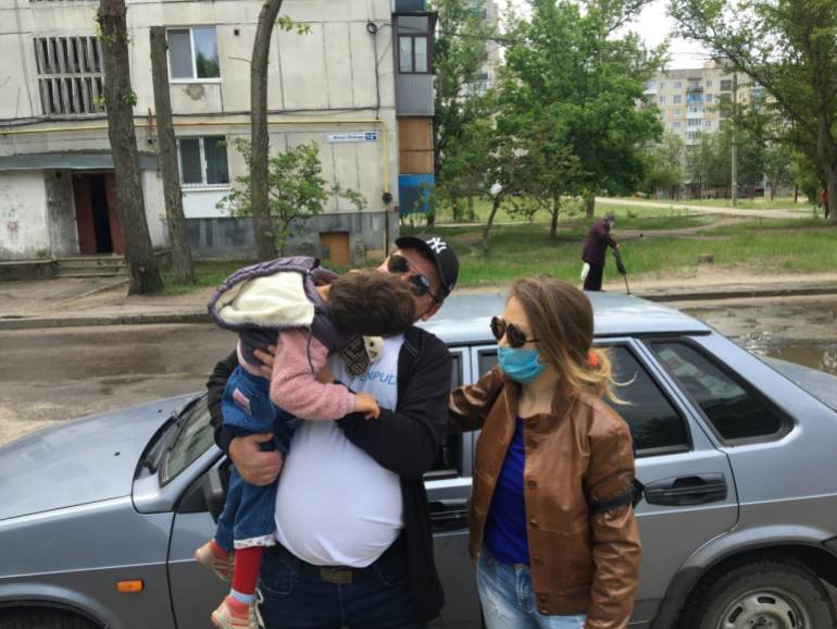 Alexey Kvitkovskiy carrying a child with a woman standing next to him with a car behind them.