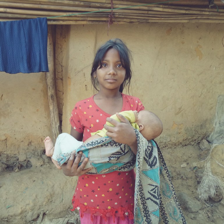 10-year-old Nur Akter, in a red dress, holds her four month old sister, wrapped in a cloth, outside their house in Kutupalong camp