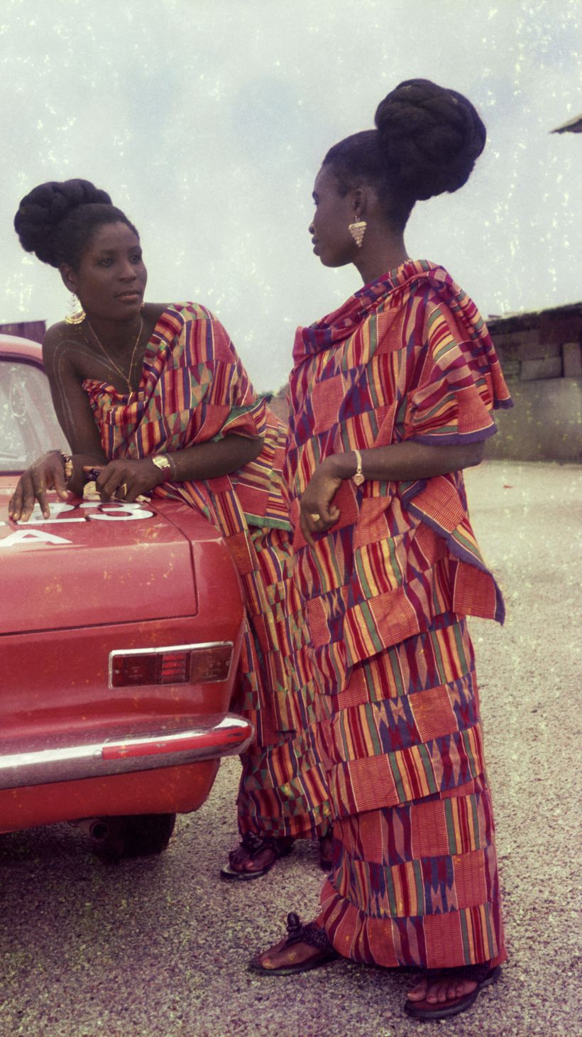 Two women dressed for a church celebration stand next to a red car