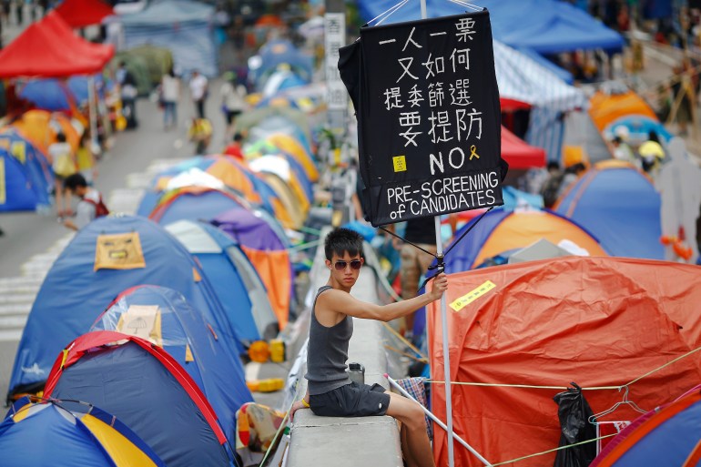 A pro-democracy protester holds a banner with tents set up on the street around him. 