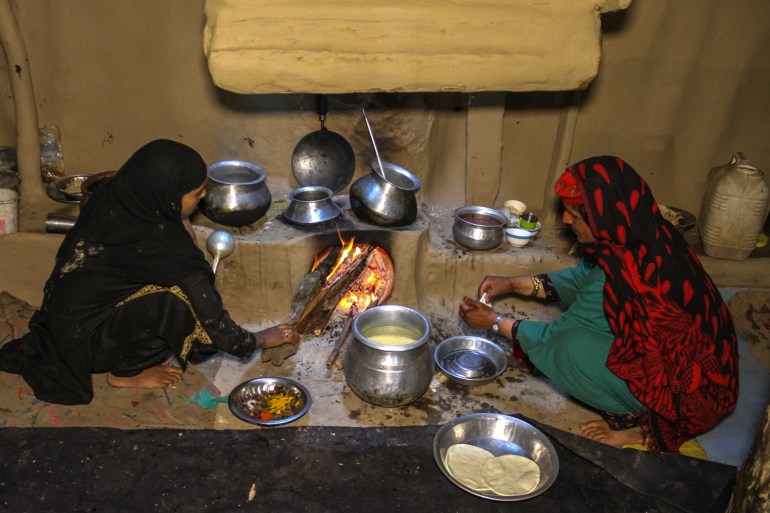 Zulaikha is seen in her kitchen at the hearth with another woman. ZUlaikha is patting out kaladi while the other woman stokes the fire