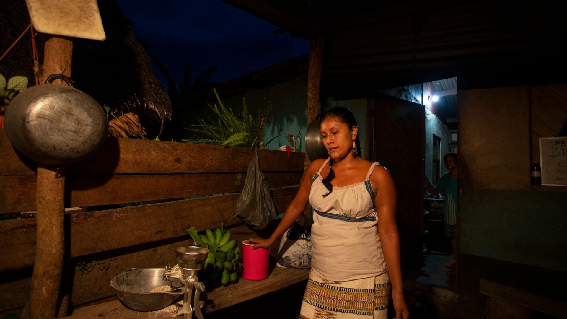 A photo of Cecilia Leyva, 35, leaning on a wooden table with a woman standing in the back near an open door.