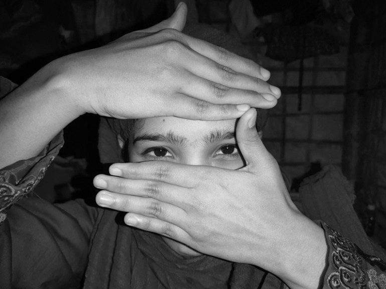 A black and white photo of a young Rohingya woman with her hands covering her mouth and forehead so only her eyes are visible