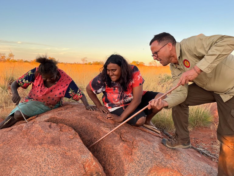 Gregory Andrew pokes a stick into the crevice of a rock while two Aboriginal women watch.