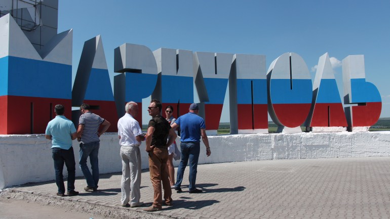 People stand near the name of the city of Mariupol written in Russian and painted in the colours of the Russian national flag during celebration of Russia Day in Mariupol, June 12, 2022