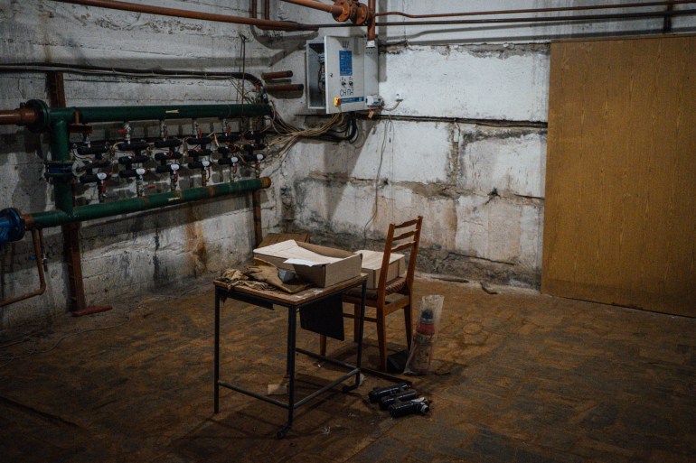 A photo of a table and chair in a dirty, rusty room with a wall of a mechanical device in front of the table and chair and a box of papers and gloves and cloth on the side of it on top of the the table.