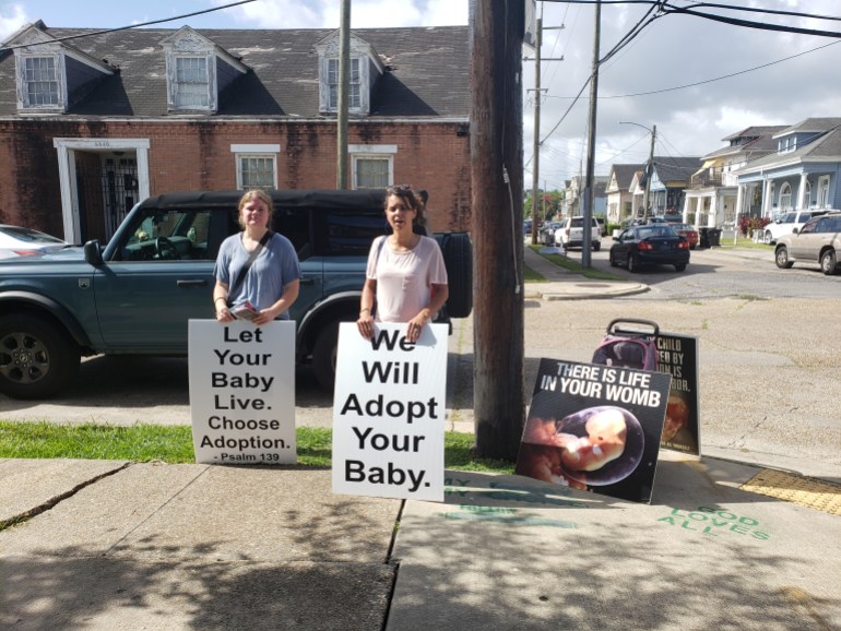 A photo of two protestors holding posters, the one on the left says "Let your baby live. Choose adoption. - Psalm, 139" and the one on the right says "We will adopt your baby."