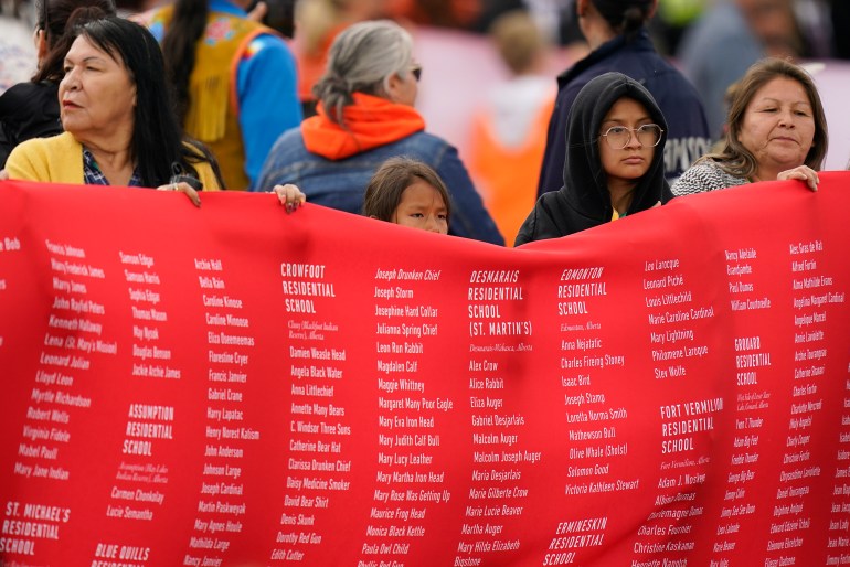 A photo of indigenous people holding up a large red banner that has a long list of names.