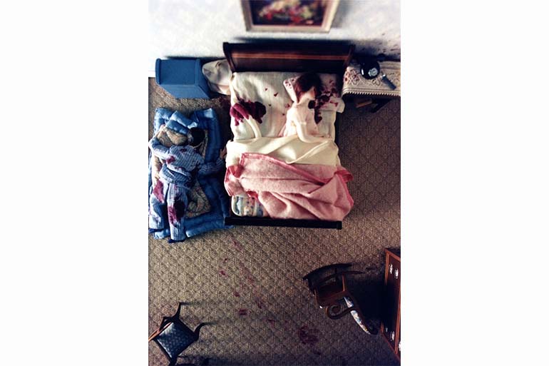 A photo of a diorama of a doll in a bed with blood on the other side of the bed and around the pillow it's lying in and a doll lying on the floor on the side of the bed with blood on it's pyjamas.