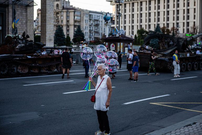 An elderly woman sells glowing balloons next to a destroyed Russian military equipment in Kyiv, ahead of Ukraine's Independence Day.