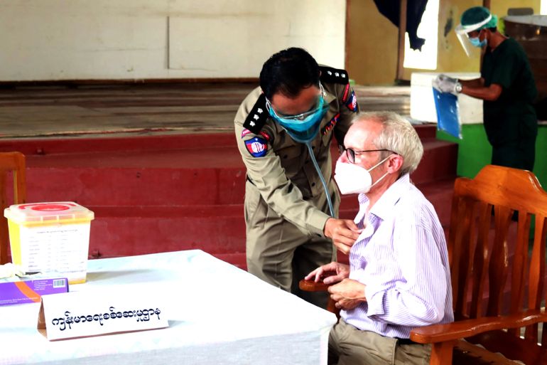Sean Turnell, wearing a face mask and white shirt, pictured by the military junta sitting at a table being given a COVID-19 vaccination