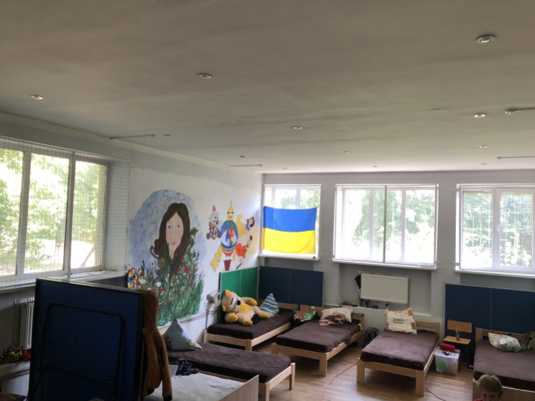 A photo of a bedroom with several small beds and a painting on one of the walls with a Ukrainian flag on one of the windows.