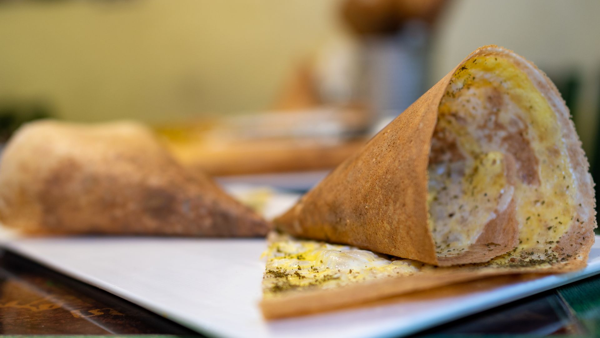 In focus is one half of a double cone of crisp regag bread. We can see the thin egg filling and a sprinkle of green zaatar