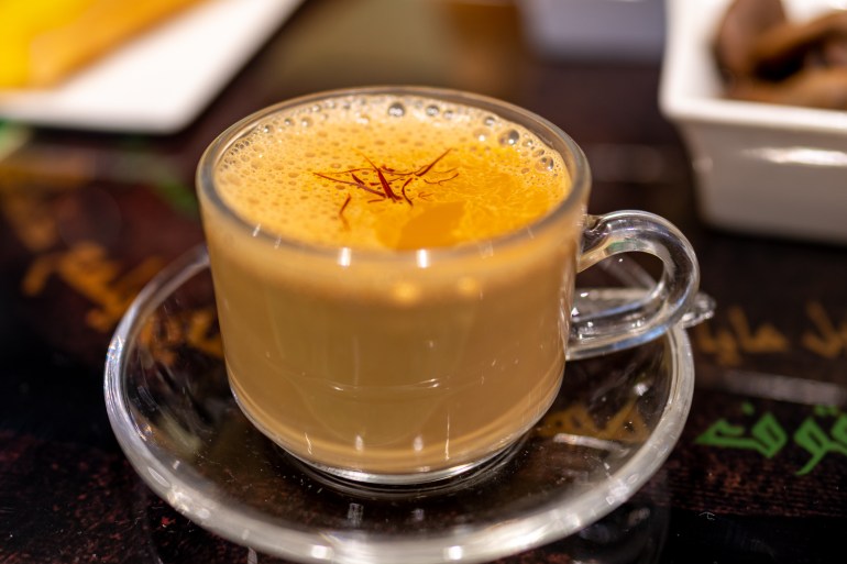 A glass cup of milky tea sits on its glass saucer. There are strands of saffron on top