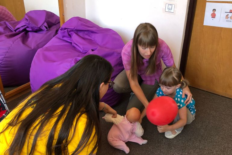 A photo of Viktoria Plyush holding her daughter while her daughter holds a balloon and reaches for a baby doll a woman in front of her is holding.