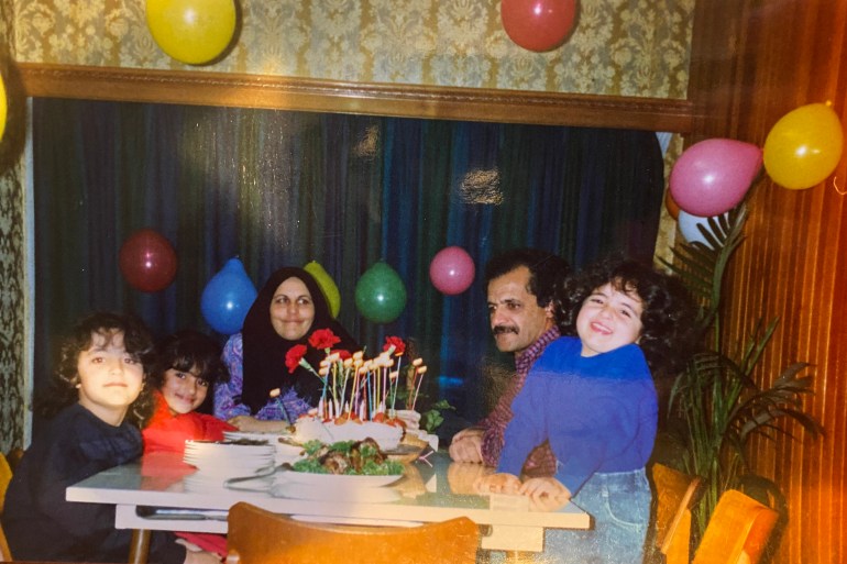 A young family of mother, father, and three daughters gathered around a table with cake, kebab and naan, and a stack of plates. The room is decorated with colourful balloons