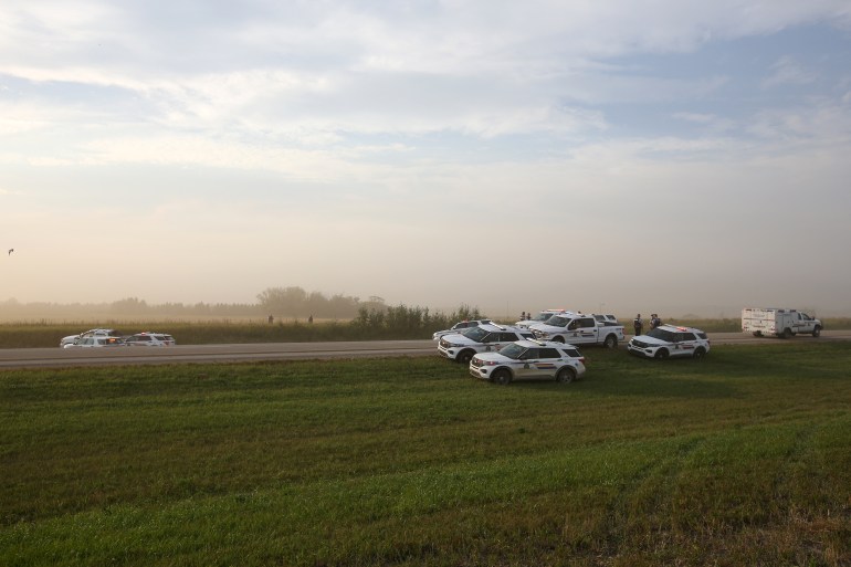 Police cars on the verge around a white truck driven by Myles Sanderson against a backdrop of flat fields and trees