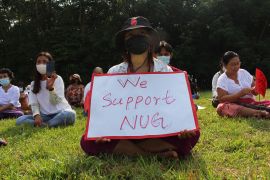 A protester in Myanmar sits on the grass with a sign reading 'We support NUG'.