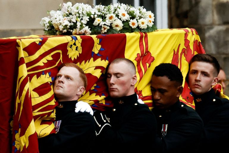 Four pallbearers carry the coffin of Queen Elizabeth II draped in a flag and with a wreath of white flowers from her Balmoral Estate on top