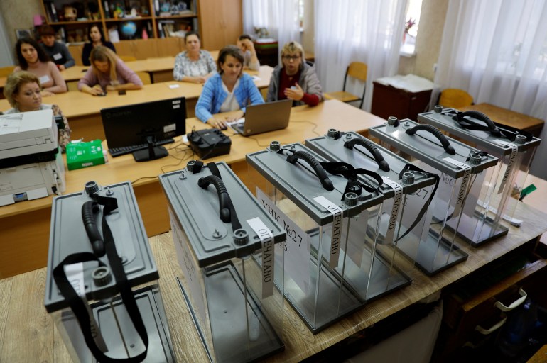 A series of clear ballot boxes laid out on a table at a polling station with members of the local electoral commission in the self-proclaimed Donetsk People's Republic sitting behind.