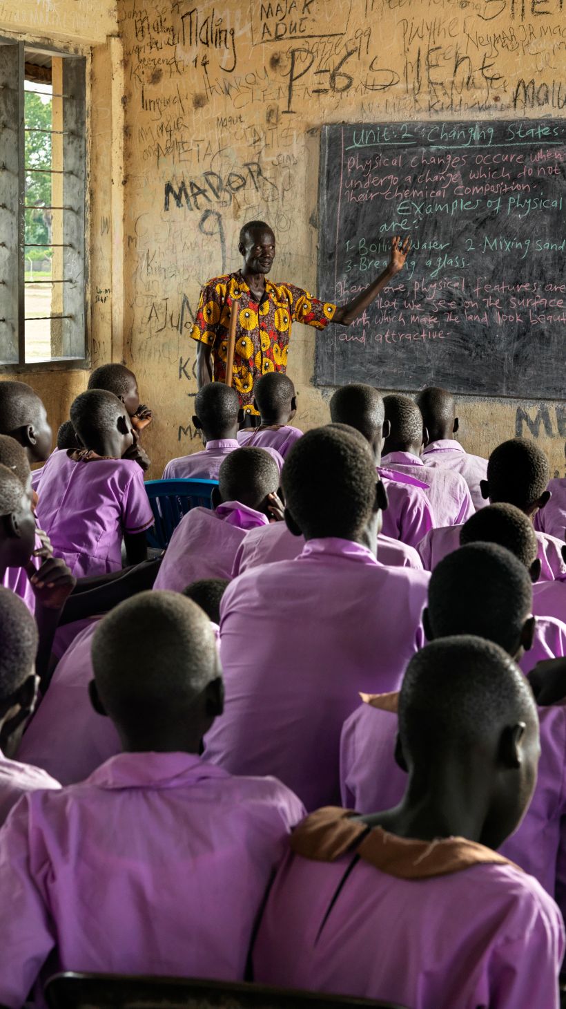 A photo of a classroom of students with a teacher in the front pointing to something on a blackboard.