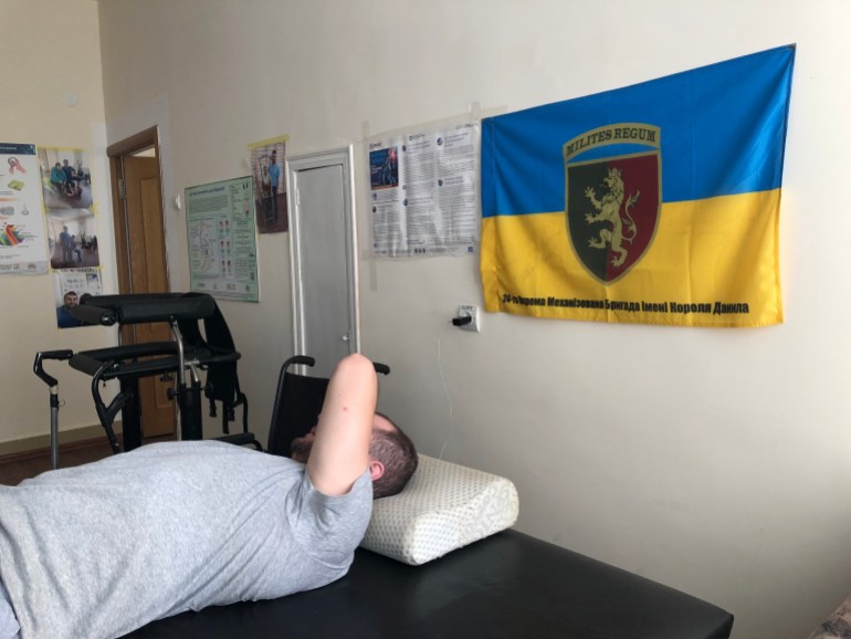 A photo of a man lying down with a flag on the wall behind him and he has his arm over his face.