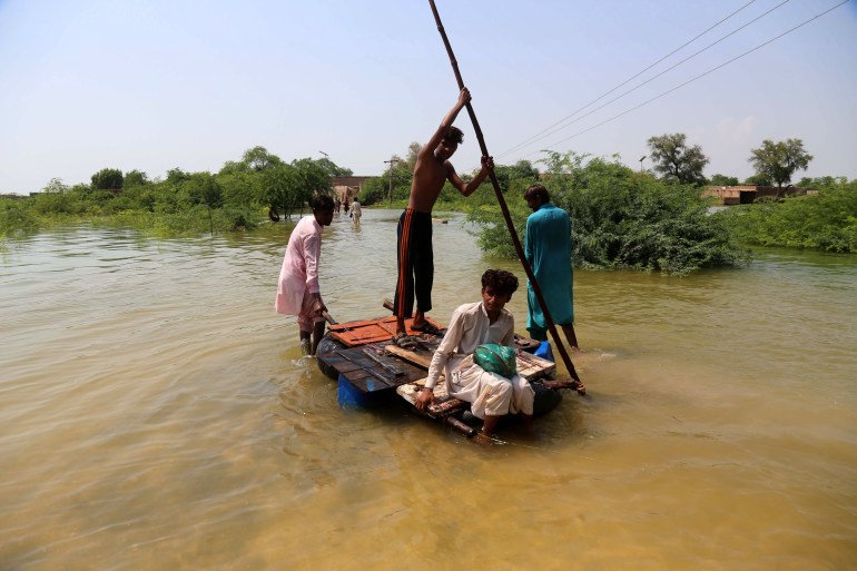 Several people sit a wooden raft on flood waters, as a man pushes the raft forward using a large stick