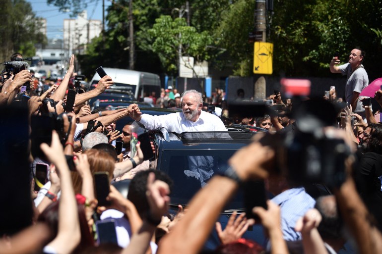 Lula in an open top car driving along a road lined by supporters trying to take pictures
