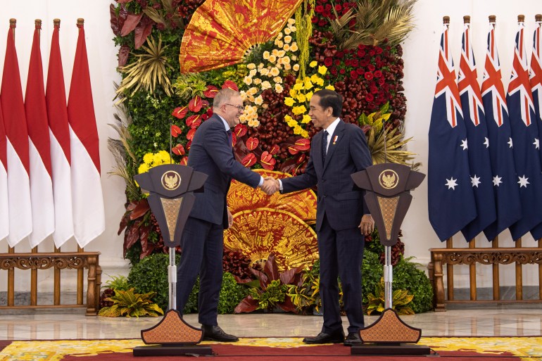 Indonesian president Joko Widodo and Australian prime minister Anthony Albanese shake hands and smile against a backdrop of flowers and their respective countries' flags