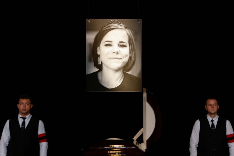 Memorial service for Darya Dugina with her coffin flanked by two men and a black and white portrait of her behind.