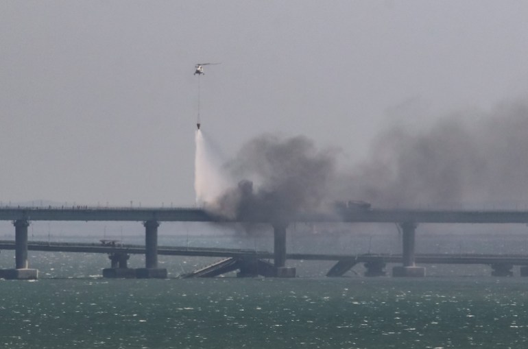A helicopter drops water to extinguish fuel tanks ablaze next to damaged sections of the Kerch bridge.