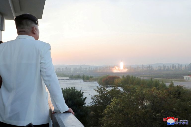 Kim Jong Un in a white jacket with his back to the camera watches a missile launch from a balcony.
