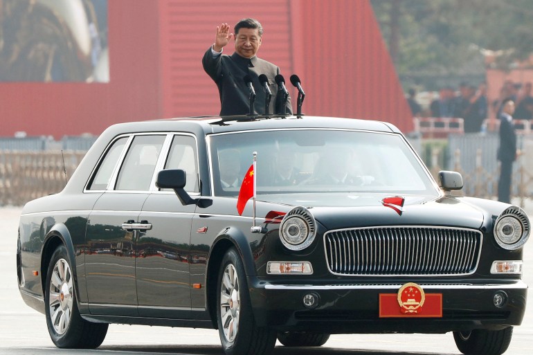 Xi Jinping standing in a classic black car with Chinese flags on each wing, stands and reviews the troops in 