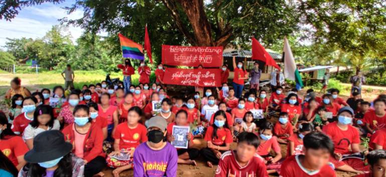 Women in red t-shirts sitting beneath a tree with flags and placards as part of a peaceful protest.