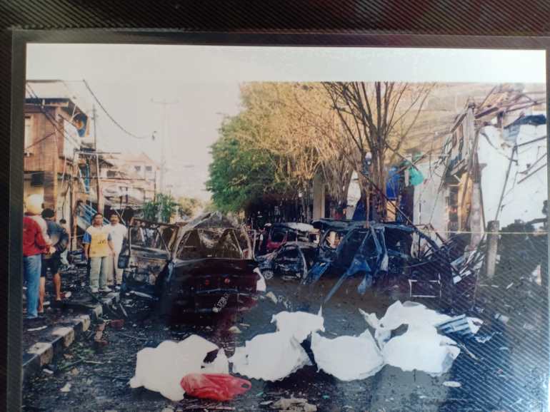 A photo showing Sadrjono's destroyed car and debris after the 2002 Bali Bombings with bodies covered by white sheets in front