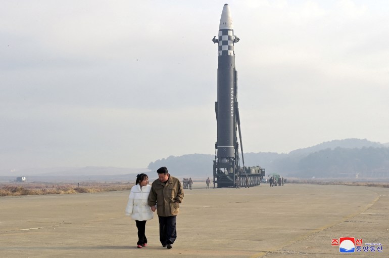 North Korean leader Kim Jong Un walks away from an intercontinental ballistic missile while holding hands with his daughter. He is looking down at her and she up at him and they seem to be talking. She is wearing what looks to be a white puffer jacket and black pants