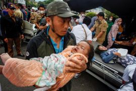 A man cradles an injured infant following the earthquake. The baby has blood on their face and the man looks worried and dazed.