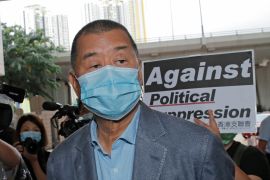 Jimmy Lai, wearing a grey suit and a medical face mask, arriving at court in 2020. Supporters are in the background holding placards with one reading 'Against political suppression'.