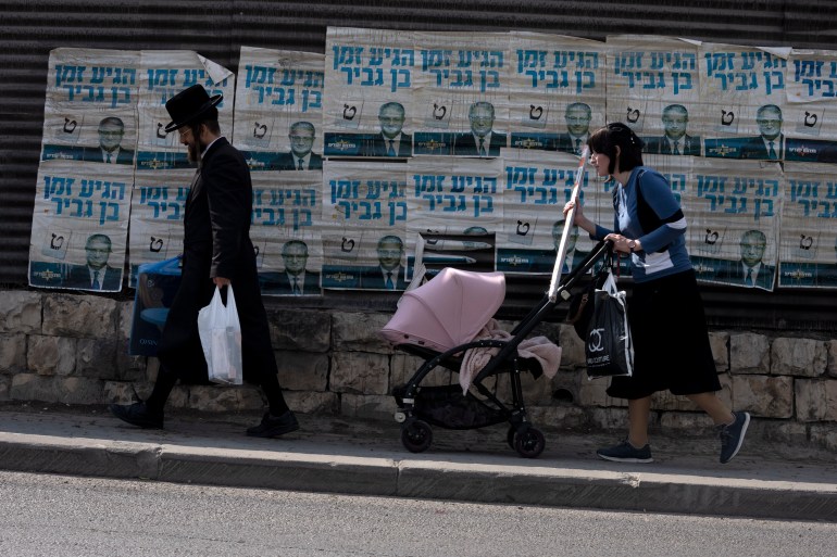 An ultra-Orthodox Jewish couple with a baby in a pushchair walk past election campaign posters for Israeli legislator Itamar Ben Gvir.