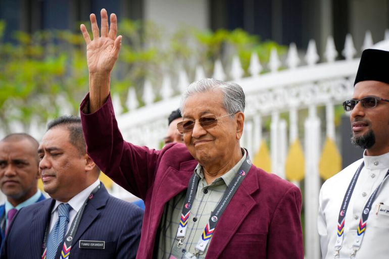 Former prime minister wearing maroon jacket and sunglasses waves, smiling