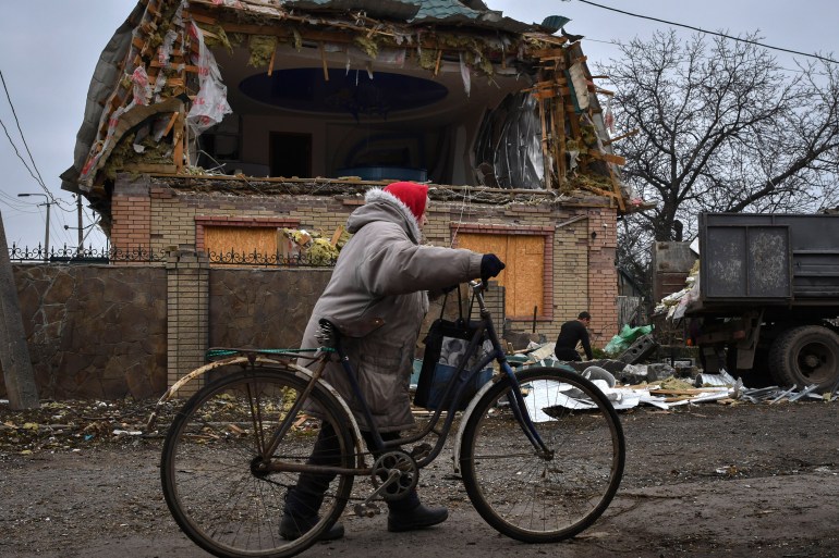 A woman passes with a bicycle as a local resident works to clean the debris from a damaged house after Russian shelling