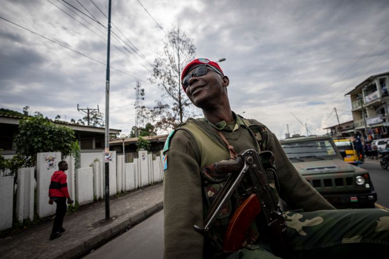 A Congolese soldier escorting a convoy of Kenya's defence forces, sitting atop car wearing red knitted hat and green uniform, leaning back and smiling