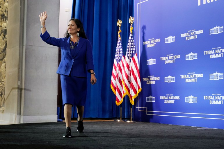 Deb Haaland waves from the stage as she walks forward, with two American flags behind her